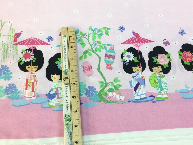 May include: Pink fabric with a repeating pattern of cartoon geishas, parasols, and lanterns. The geishas are in traditional attire, with black hair and floral accents. The fabric is measured with a wooden ruler.