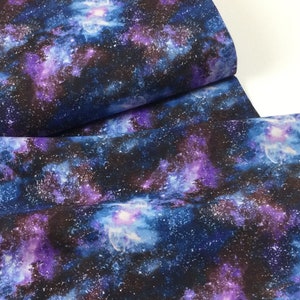 May include: A fabric with a galaxy design. The fabric features a dark blue background with swirling patterns of purple, white, and light blue, resembling stars and nebulae. The fabric is folded, showing its texture and pattern.
