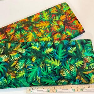 May include: Two fabric pieces with vibrant tropical leaf patterns. One features green, yellow, and orange leaves on a dark background. The other showcases lush green leaves with hints of blue and yellow. A ruler is placed at the bottom.