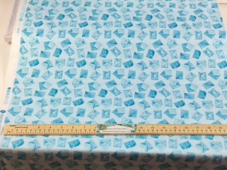 May include: Light blue fabric with a watercolor pattern of scattered envelopes in varying shades of blue. A wooden ruler with inch measurements is placed across the fabric. The fabric is likely for sewing or crafting projects.