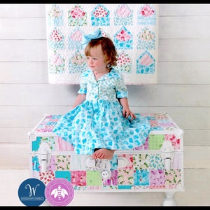 May include: A young child wearing a blue floral dress and a matching bow sits on a patchwork trunk. The trunk is decorated with various floral patterns and colors. A quilt with floral envelope designs hangs in the background.