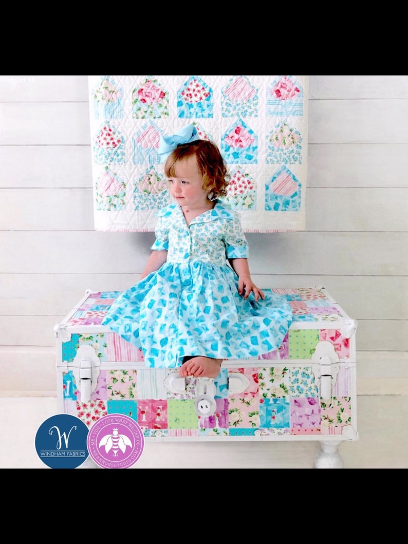 May include: A young child wearing a blue floral dress and a matching bow sits on a patchwork trunk. The trunk is decorated with various floral patterns and colors. A quilt with floral envelope designs hangs in the background.