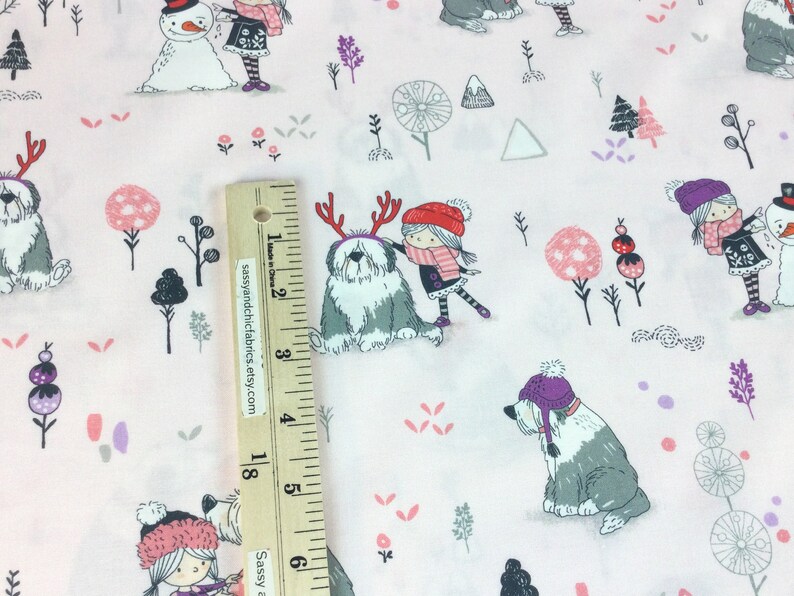 May include: Light pink fabric with a repeating pattern of Old English Sheepdogs, girls, snowmen, and trees. The dogs wear antler headbands and hats. The girls wear hats and scarves. The fabric is decorated with pink, purple, and gray elements.