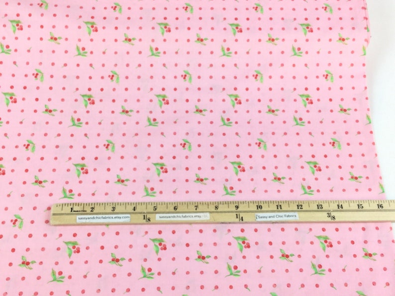 Puede incluir: Tela rosa con un patr&oacute;n repetido de cerezas rojas y hojas verdes, intercaladas con lunares rojos. Una regla con medidas en pulgadas est&aacute; colocada sobre la tela. El texto "sassyandchicfabrics.etsy.com" es visible en la regla.