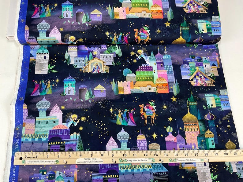 Puede incluir: Tela con un dise&ntilde;o colorido y caprichoso que presenta una escena de la natividad, edificios, estrellas y figuras sobre un fondo azul oscuro. La tela tiene una regla con medidas en cent&iacute;metros.