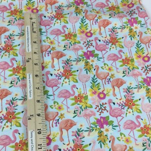 May include: Light blue fabric with a repeating pattern of pink and orange flamingos, yellow and orange flowers, and green leaves. A wooden ruler is placed on the fabric. The text "Sassy and Chic Fabrics" is visible.