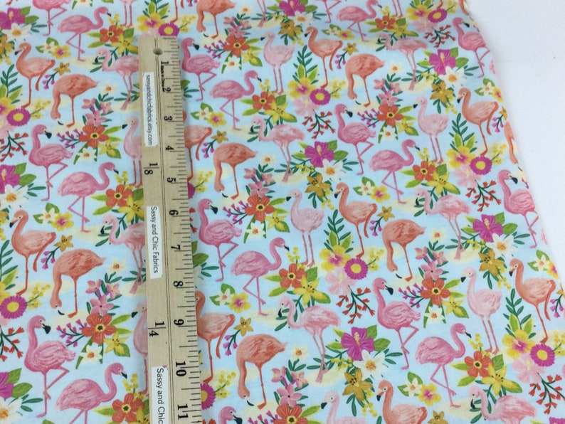 May include: Light blue fabric with a repeating pattern of pink and orange flamingos, yellow and orange flowers, and green leaves. A wooden ruler is placed on the fabric. The text "Sassy and Chic Fabrics" is visible.