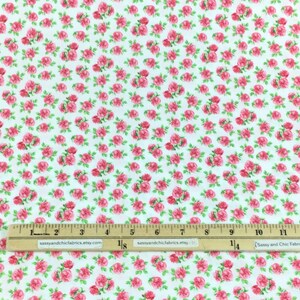 May include: White fabric covered in a repeating pattern of small pink roses with green leaves. A wooden ruler with inch measurements is placed across the fabric. The fabric is likely used for sewing or crafting.