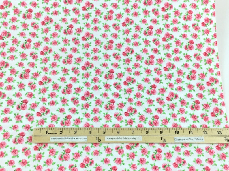 May include: White fabric covered in a repeating pattern of small pink roses with green leaves. A wooden ruler with inch measurements is placed across the fabric. The fabric is likely used for sewing or crafting.