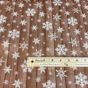 Pu&ograve; includere: Tessuto marrone con motivo a venature di legno, ornato da fiocchi di neve bianchi di varie dimensioni. &Egrave; visibile un righello con pollici e il testo "sassyandchicfabrics.etsy.com". Adatto per lavori artigianali e decorazioni invernali.