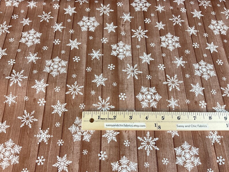 Pu&ograve; includere: Tessuto marrone con motivo a venature di legno, ornato da fiocchi di neve bianchi di varie dimensioni. &Egrave; visibile un righello con pollici e il testo "sassyandchicfabrics.etsy.com". Adatto per lavori artigianali e decorazioni invernali.