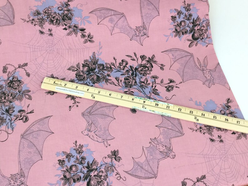 Puede incluir: Tela rosa con un patr&oacute;n repetido de murci&eacute;lagos, telara&ntilde;as y ramos florales en negro y azul claro. Una regla de madera se coloca en diagonal sobre la tela, midiendo hasta 43 cm.