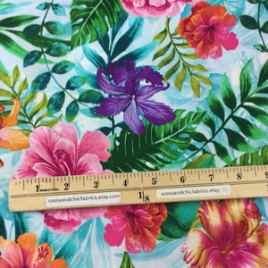 May include: Fabric with a tropical floral pattern featuring large pink, orange, and purple flowers, along with green leaves, set against a light blue background. A wooden ruler is placed across the fabric. The fabric is likely used for sewing or crafting projects.