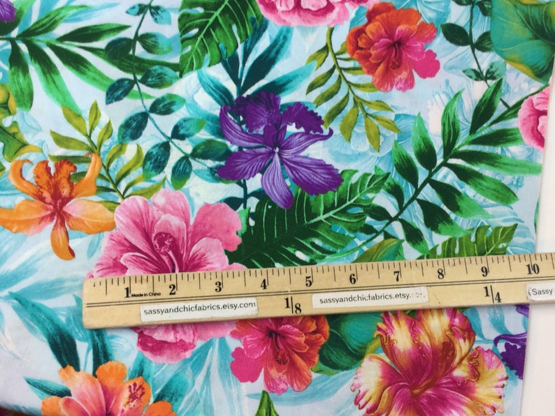 May include: Fabric with a tropical floral pattern featuring large pink, orange, and purple flowers, along with green leaves, set against a light blue background. A wooden ruler is placed across the fabric. The fabric is likely used for sewing or crafting projects.