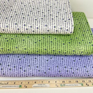 May include: Three folded bolts of fabric in white, light green, and light purple, each featuring a repeating pattern of small lavender flowers and text including phrases like A Passion for Purple and Be Kind, with a 16-inch wooden ruler below.