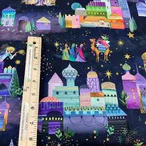 Puede incluir: Tela con una ilustraci&oacute;n caprichosa de un paisaje urbano nocturno con edificios coloridos, estrellas y figuras. El dise&ntilde;o incluye un camello y tres figuras, posiblemente representando un tema religioso. Una regla de madera est&aacute; colocada sobre la tela.