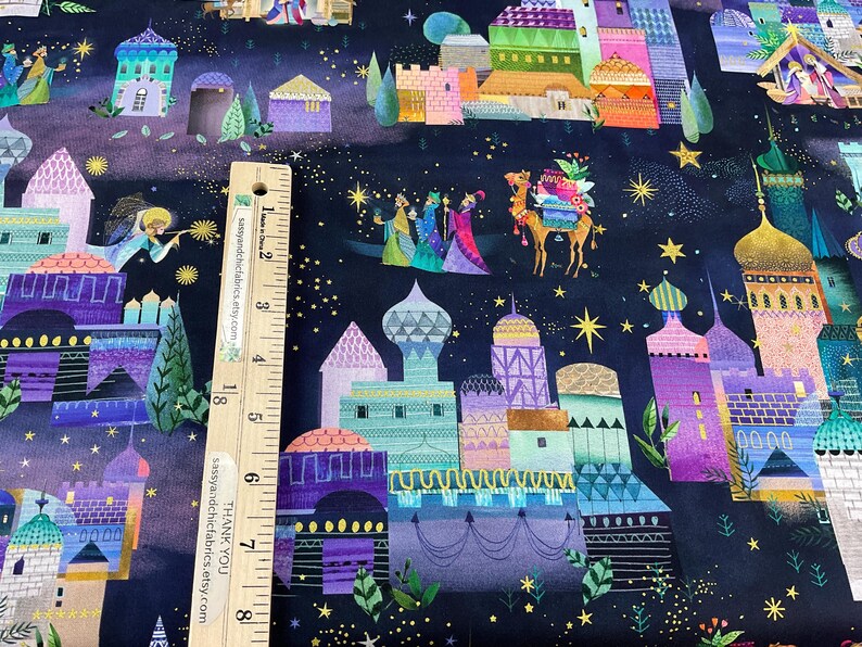 Puede incluir: Tela con una ilustraci&oacute;n caprichosa de un paisaje urbano nocturno con edificios coloridos, estrellas y figuras. El dise&ntilde;o incluye un camello y tres figuras, posiblemente representando un tema religioso. Una regla de madera est&aacute; colocada sobre la tela.