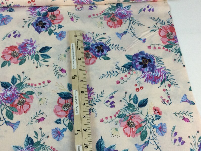 May include: Cream-coloured floral fabric with a pattern of pink, purple, and blue flowers and green leaves. A ruler is placed on the fabric, showing measurements in inches.