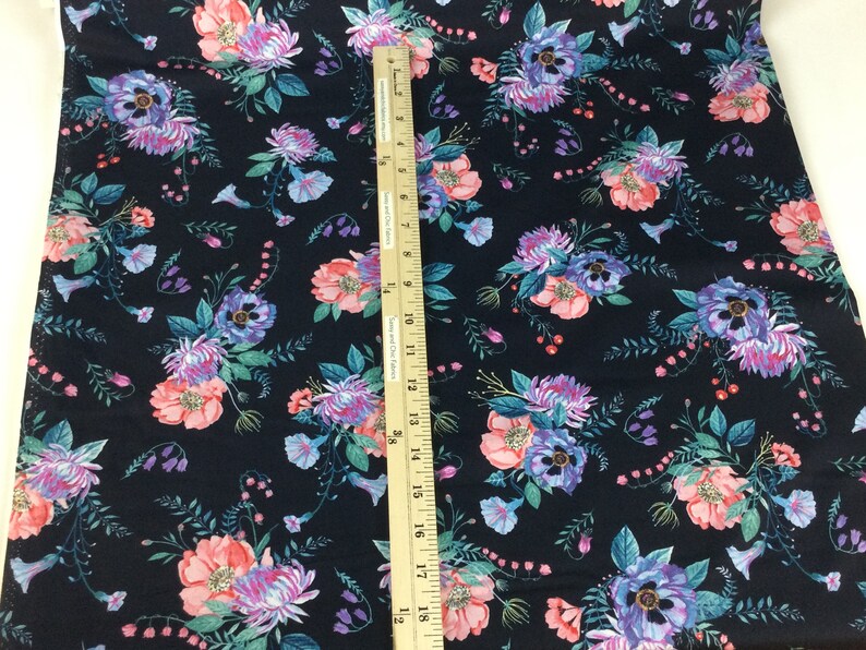 May include: Dark navy fabric with a floral pattern featuring pink, purple, and blue flowers with green leaves. A wooden ruler is placed on the fabric, showing measurements.