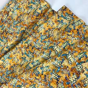 May include: Fabric with a repeating pattern of sea turtles in shades of blue, tan, and brown. The turtles are detailed with textured shells and flippers, set against a mottled background. The fabric is folded, showing multiple layers.