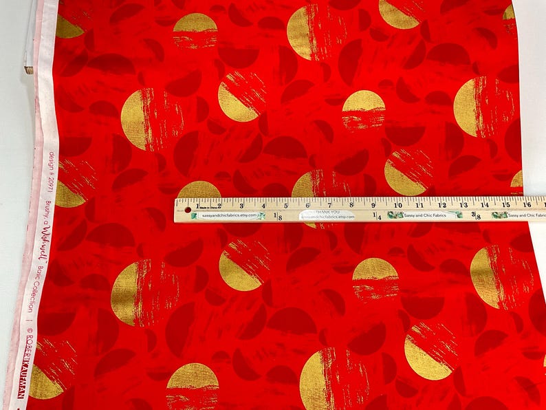 May include: Red fabric with a repeating pattern of gold half-circles and brushstroke textures. The design includes smaller, darker red leaf shapes. A wooden ruler is placed across the fabric, measuring up to 16 inches.