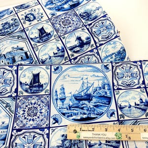 May include: White fabric with a blue Delftware-style pattern. The design features nautical scenes, windmills, and floral motifs within square and circular frames. A ruler is visible, indicating the fabric's dimensions.