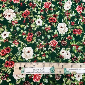 K&ouml;nnte beinhalten: Stoff mit dunkelgr&uuml;nem Hintergrund, der ein Muster aus wei&szlig;en und roten Rosen, Stechpalmenbl&auml;ttern und roten Beeren zeigt. Kleine Tannenzapfen sind ebenfalls &uuml;ber das Design verstreut. Ein Lineal liegt auf dem Stoff.