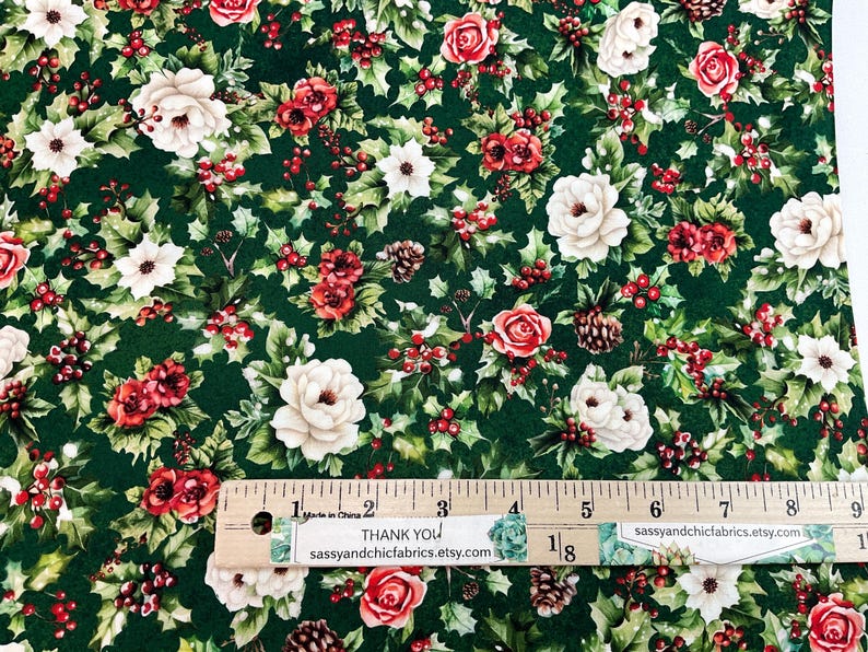 K&ouml;nnte beinhalten: Stoff mit dunkelgr&uuml;nem Hintergrund, der ein Muster aus wei&szlig;en und roten Rosen, Stechpalmenbl&auml;ttern und roten Beeren zeigt. Kleine Tannenzapfen sind ebenfalls &uuml;ber das Design verstreut. Ein Lineal liegt auf dem Stoff.
