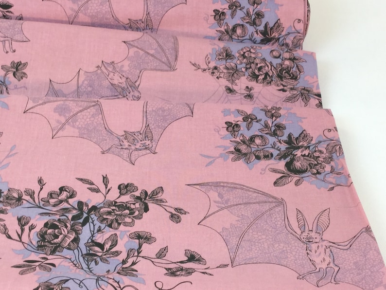 Puede incluir: Tela rosa con un patr&oacute;n repetido de murci&eacute;lagos y dise&ntilde;os florales en negro y morado claro. Los murci&eacute;lagos se representan con las alas extendidas, y las flores incluyen rosas y otras flores. La tela est&aacute; doblada, mostrando m&uacute;ltiples capas.