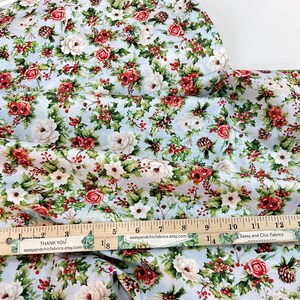 K&ouml;nnte beinhalten: Blumenstoff mit hellblauem Hintergrund, mit roten Rosen, wei&szlig;en Blumen, gr&uuml;nen Bl&auml;ttern und Tannenzapfen. Ein Holzlineal zeigt Messungen bis zu 38 cm. Der Stoff stammt von Sassy and Chic Fabrics.