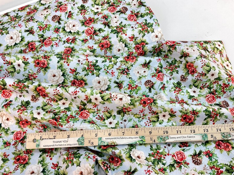K&ouml;nnte beinhalten: Blumenstoff mit hellblauem Hintergrund, mit roten Rosen, wei&szlig;en Blumen, gr&uuml;nen Bl&auml;ttern und Tannenzapfen. Ein Holzlineal zeigt Messungen bis zu 38 cm. Der Stoff stammt von Sassy and Chic Fabrics.