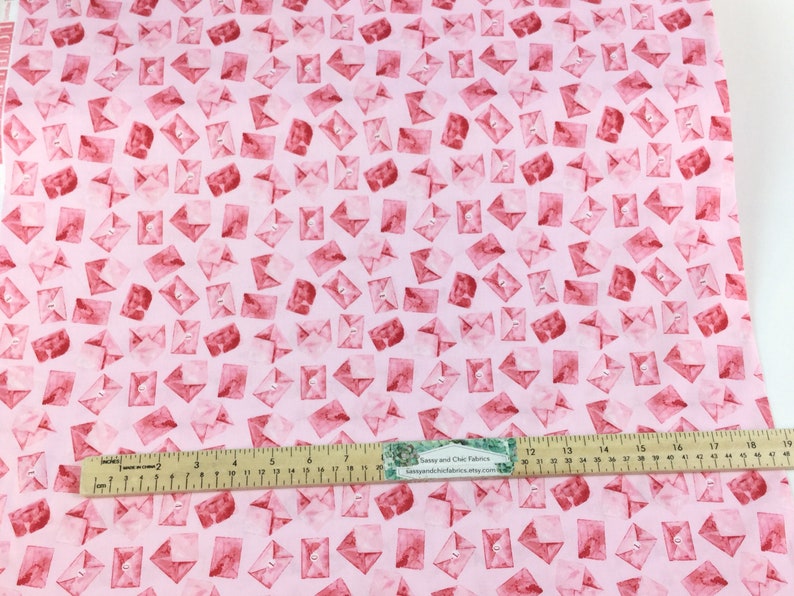 May include: Pink fabric with a repeating pattern of red watercolor envelopes. Each envelope has a small button detail. A ruler with inch measurements is placed on the fabric.