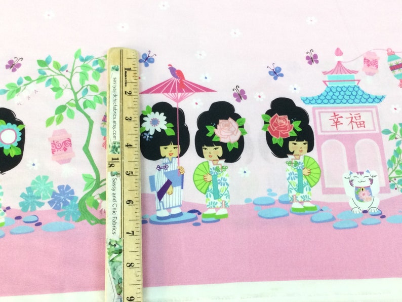 May include: Pink fabric with a Japanese-themed design. Features illustrations of geishas, pagodas, and lucky cats. The fabric includes a ruler for scale, showing the pattern's size.