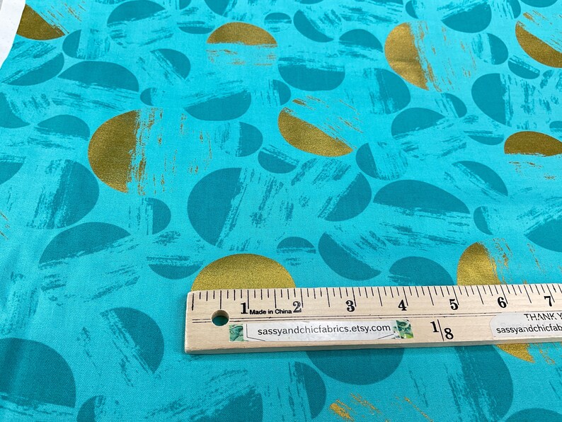 May include: Teal fabric with a pattern of gold and teal half-circles and brushstrokes. A wooden ruler with inch measurements is placed on the fabric. The text "Made in China" and "sassyandchicfabrics.etsy.com" are visible.