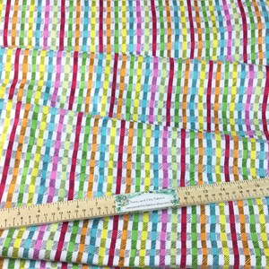 May include: Colorful fabric with a vertical striped pattern in red, orange, yellow, green, blue, and pink. The fabric is white with a grid-like design. A wooden ruler with inch measurements is placed on the fabric.