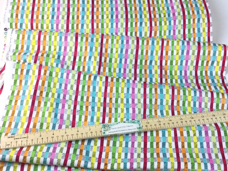 May include: Colorful fabric with a vertical striped pattern in red, orange, yellow, green, blue, and pink. The fabric is white with a grid-like design. A wooden ruler with inch measurements is placed on the fabric.