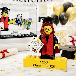May include: A graduation-themed Lego display featuring two minifigures in graduation attire, holding diplomas. One is in a clear case, the other on a yellow base with the name "ANNA Class of 2026". The banner reads "CONGRATULATIONS".