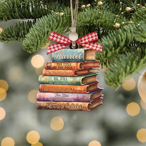 May include: A wooden Christmas ornament shaped like a stack of books. The top book has the name "Harriet" on it. Each book has a title, such as "The Title of the Book 1". The ornament is decorated with a red and white bow.