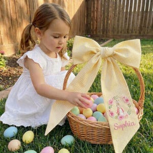 May include: A wicker Easter basket with a large yellow gingham bow and a personalized ribbon. The basket is filled with pastel-colored eggs. The ribbon features a floral wreath with a bunny and the name "Sophia".