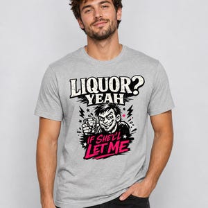 Peut inclure: T-shirt gris clair avec l'inscription "LIQUOR? YEAH IF SHE'LL LET ME". Le motif présente un personnage de dessin animé souriant tenant un verre, avec des éclairs et des étoiles.
