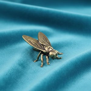 May include: A bronze fly ornament with detailed wings and legs. The insect sculpture is set against a teal fabric background, highlighting its intricate design and metallic finish. The fly is a decorative item.