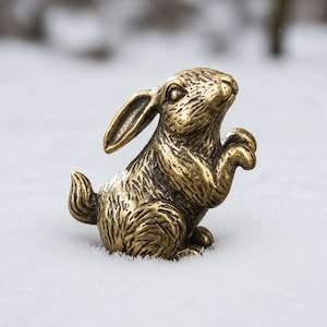May include: A small, metallic, gold-toned brass figurine of a rabbit in a seated, upright position. The rabbit has detailed, textured fur markings, one long ear pointing upward, and its front paws are raised near its chest area.