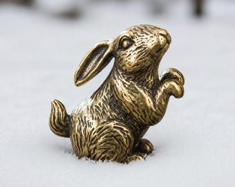 May include: A small, metallic, gold-toned brass figurine of a rabbit in a seated, upright position. The rabbit has detailed, textured fur markings, one long ear pointing upward, and its front paws are raised near its chest area.