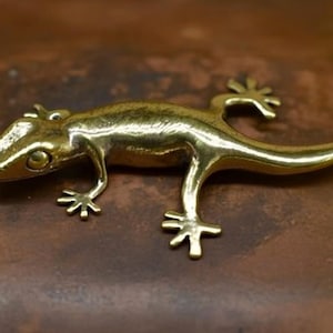 May include: A detailed, golden-colored lizard figurine. The reptile is posed with its legs spread, and its tail curved. The lizard is made of a shiny metal and is set against a brown background. The figurine is a decorative item.
