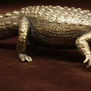 Peut inclure: Une figurine d'alligator de couleur bronze avec des écailles détaillées et une surface texturée. La sculpture est posée en position de marche, mettant en valeur ses pattes et sa queue. C'est un objet décoratif.
