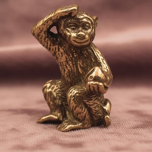 May include: A small, detailed brass monkey figurine in a seated position, with one hand raised to its head. The monkey has a textured surface and a smiling expression. The figurine is set against a soft, muted pink background.