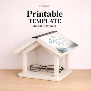 May include: A white house-shaped wooden book stand with a peaked roof design. An open book titled The Adventure Begins rests on the roof. A pair of dark-rimmed eyeglasses sits on the base inside the structure. Text above reads ceramixis Printable TEMPLATE digital download.