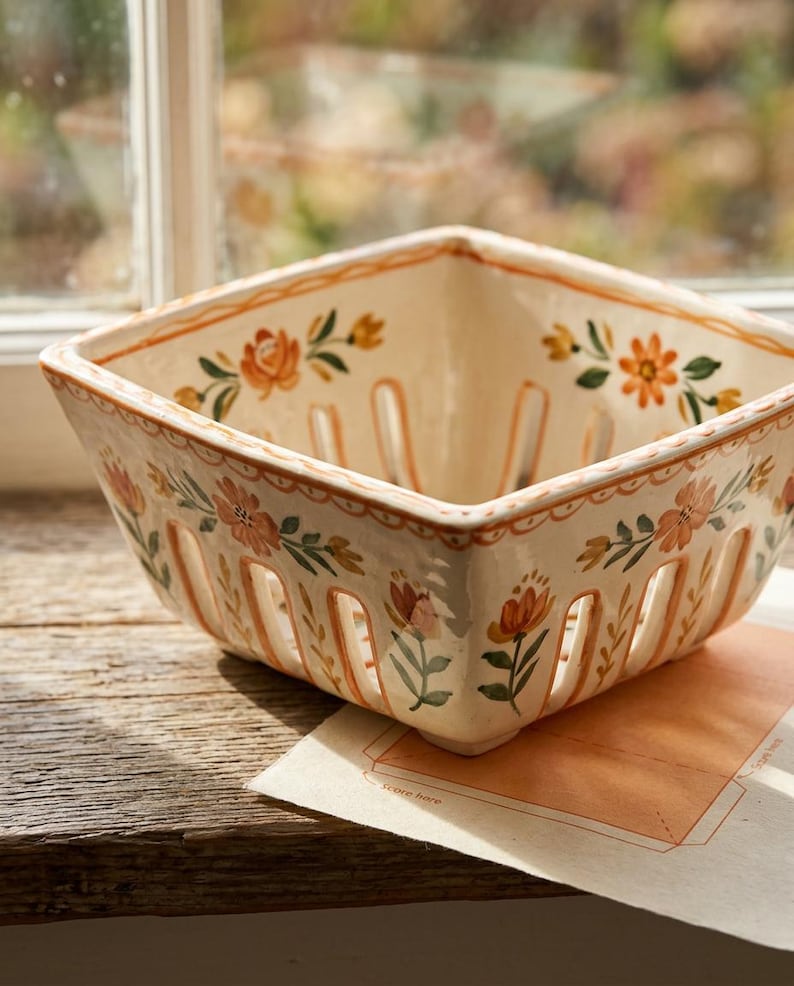 May include: Cream-colored ceramic berry basket with a square shape and slotted sides. The basket is decorated with orange and pink floral patterns and green leaves. It sits on a wooden surface near a window.