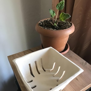 May include: A white square ceramic planter with cut-out slots sits on a wooden surface. A terracotta pot with a small plant is in the background. The planter is empty and ready for use.
