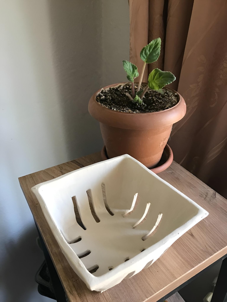 May include: A white square ceramic planter with cut-out slots sits on a wooden surface. A terracotta pot with a small plant is in the background. The planter is empty and ready for use.
